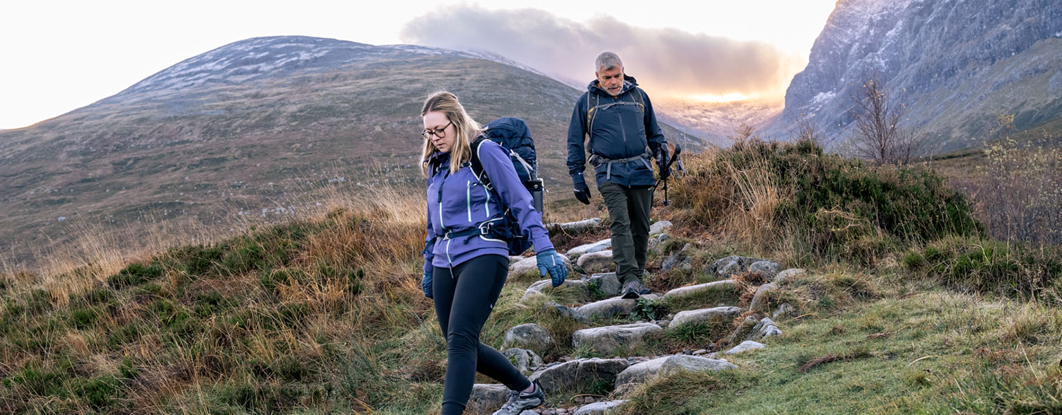 Two people are walking down on a path, wearing outdoor kit. 