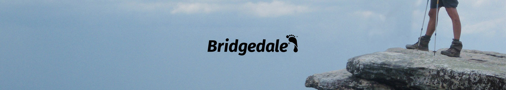 A man's leg with walking poles standing on a rock on the left with the blue sky on the right, with Bridgedale brand logo in the middle