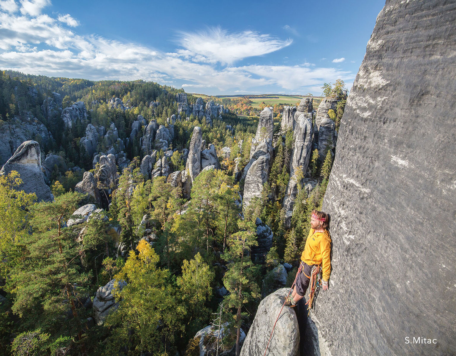 A woman is almost the top of a tall rock, wearing Patagonia gear and clinbing shoes.