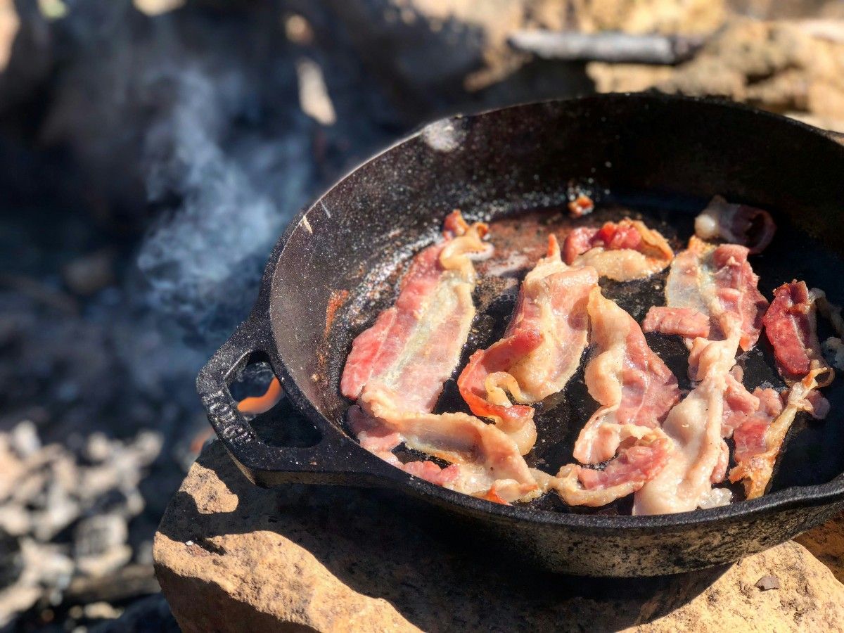 A person frying bacon over a campfire