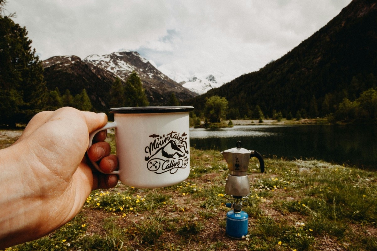 An image of a person holding their mug, with their camp kettle in the background, in front of a green hilly landscape