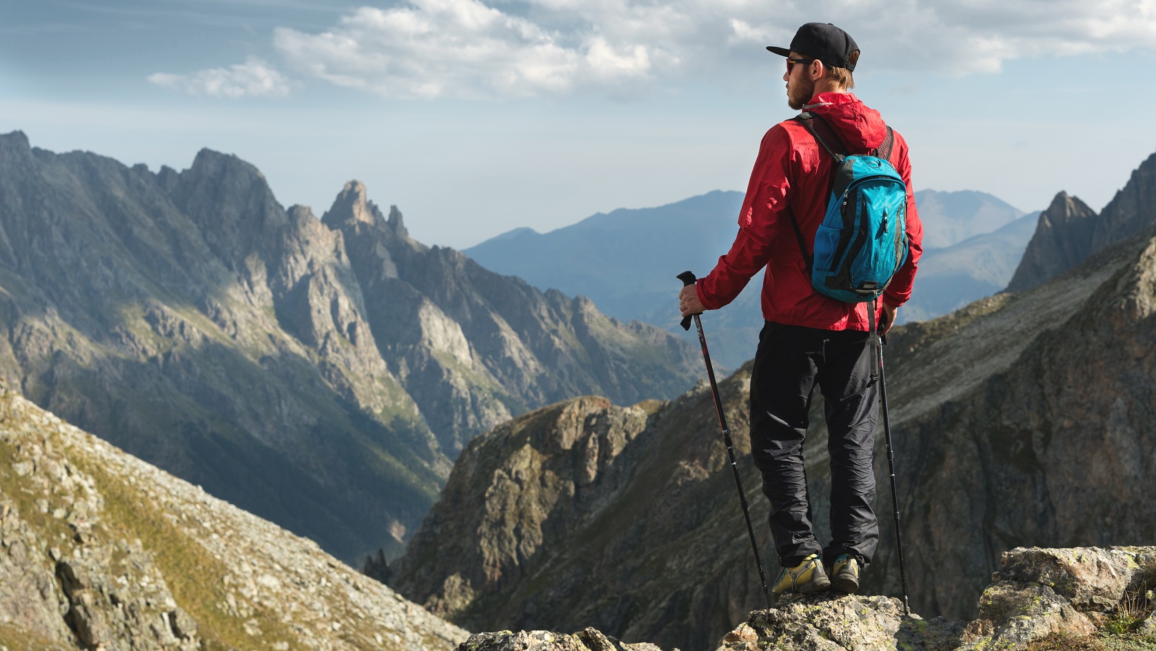 A bearded man in sunglasses and a cap with a backpack stands on top of a rock and looks into a rocky valley high in the mountains. The concept of tourism and easy trekking in the mountains outdoor.