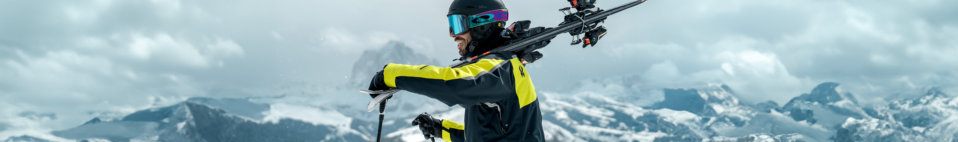 Shop Snow + Rock new season equipment. A man in yellow ski jacket carrying skies over his shoulder